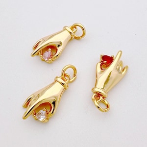 May include: Three gold hand charms with different coloured gemstones. Each charm features a hand holding a heart-shaped gemstone. The gemstones are pink, red, and clear.