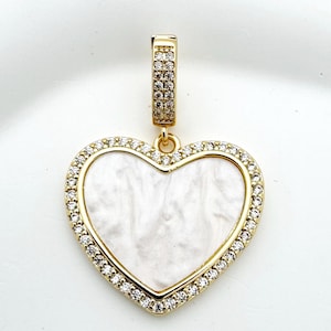 Peut inclure: Pendentif en forme de cœur doré avec un centre blanc marbré et une bordure de petits cristaux transparents. Le pendentif a une bélière dorée assortie avec des cristaux supplémentaires. La forme de cœur est un design classique.