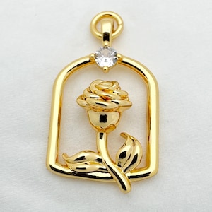 May include: A gold pendant with a rose design within a rectangular frame. The rose has detailed petals and a stem with a leaf. A small, clear gemstone is set at the top of the frame. The pendant is likely for a necklace or bracelet.