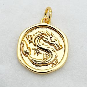May include: A gold-colored pendant with a detailed dragon design. The pendant is circular with a raised border and a small loop at the top for a chain. The dragon is intricately carved within the circle.