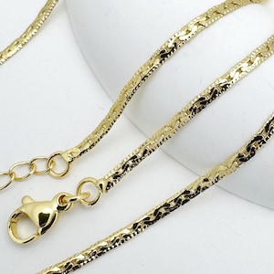 May include: A gold-colored necklace with a textured, herringbone-like chain. The necklace features a lobster clasp closure and an adjustable chain. The jewelry is displayed on a white surface.