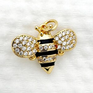 Pode incluir: Um pingente de abelha dourada com cristais transparentes e brilhantes nas asas. O corpo da abelha apresenta listras pretas e douradas. O pingente tem um pequeno laço na parte superior para prender a uma corrente ou pulseira. O fundo é um tecido branco macio.
