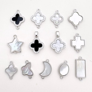 Könnte beinhalten: Sortiment an silberfarbenen Metall-Anhängern mit verschiedenen Formen und Designs, darunter ein Stern, ein Kreuz, ein Kleeblatt, ein Mond und ein Rechteck. Einige Anhänger sind mit weißen oder schwarzen Steinen verziert.