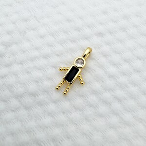 May include: A gold-colored pendant in the shape of a stick figure. The head is a clear, round stone, the body is a black rectangular stone, and the arms and legs are small gold beads. The pendant is on a white, textured surface.
