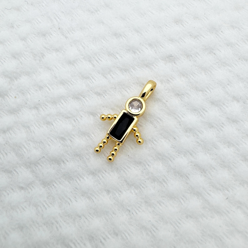 May include: A gold-colored pendant in the shape of a stick figure. The head is a clear, round stone, the body is a black rectangular stone, and the arms and legs are small gold beads. The pendant is on a white, textured surface.