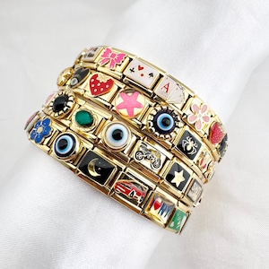 May include: Gold-toned charm bracelets with colorful enamel designs. The charms feature various symbols, including a strawberry, a star, an eye, and a playing card. The bracelets are stacked on a white surface.