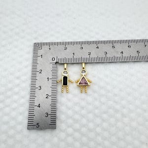 May include: Two gold-colored pendant charms in the shape of stick figures. One has a black rectangular body and the other a pink triangular body. Both have clear gemstone heads and small beaded arms and legs. A ruler is in the background.