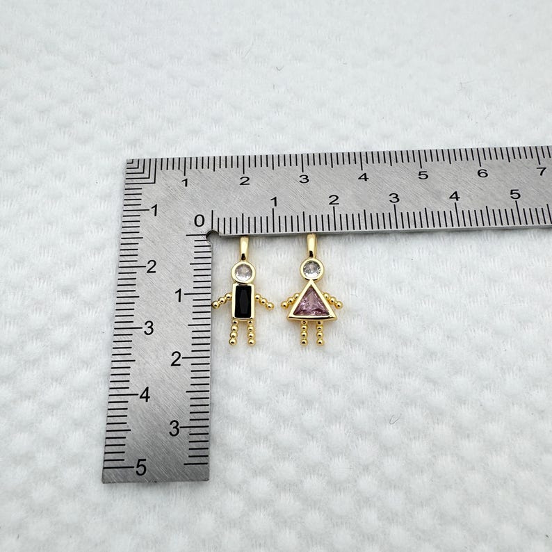 May include: Two gold-colored pendant charms in the shape of stick figures. One has a black rectangular body and the other a pink triangular body. Both have clear gemstone heads and small beaded arms and legs. A ruler is in the background.