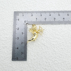 May include: A gold-colored pendant in the shape of Cupid, holding a bow and arrow. The pendant is approximately 1 inch in length. The pendant is shown next to a ruler for scale.