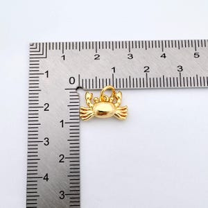 May include: Gold-colored metal crab charm pendant measuring approximately 1 inch in length. The charm is shown next to a ruler for size reference.