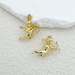 May include: Two gold-colored cherub pendants, each holding a bow and arrow. The cherubs have wings and are in a flying pose. The pendants are on a white textured surface.