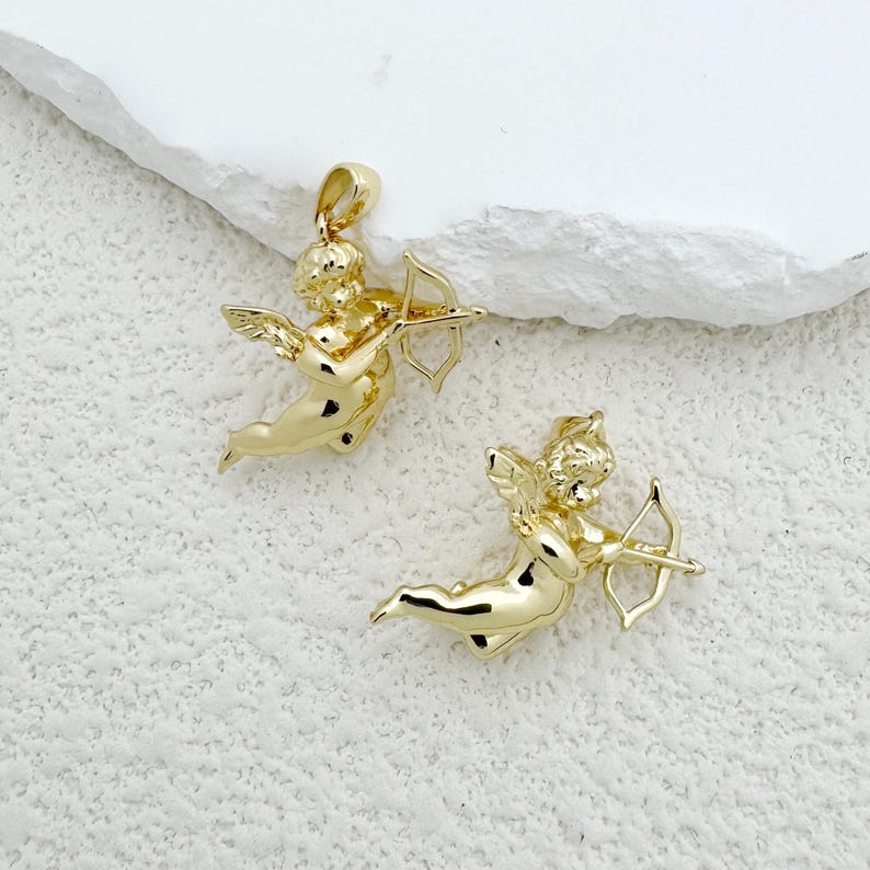 May include: Two gold-colored cherub pendants, each holding a bow and arrow. The cherubs have wings and are in a flying pose. The pendants are on a white textured surface.