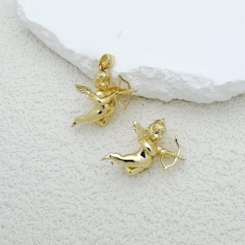 May include: Two gold-colored cupid pendants, each holding a bow and arrow. The pendants feature detailed wings and flowing hair, with a loop at the top for a chain. The background is a textured white surface.