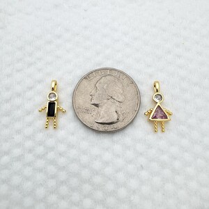 May include: Two gold-colored pendant charms in the shape of stick figures. One has a black rectangular body and the other has a pink triangular body. Both have a clear, round gem for a head and small gold beads for arms and legs.