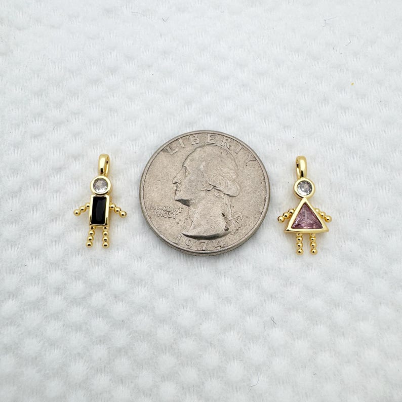 May include: Two gold-colored pendant charms in the shape of stick figures. One has a black rectangular body and the other has a pink triangular body. Both have a clear, round gem for a head and small gold beads for arms and legs.