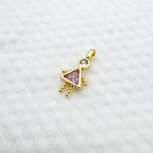 May include: A gold-colored pendant in the shape of a stylized person. The pendant features a pink triangle for the body, a clear round stone for the head, and small gold beads for the arms and legs. The pendant has a loop at the top for a chain.
