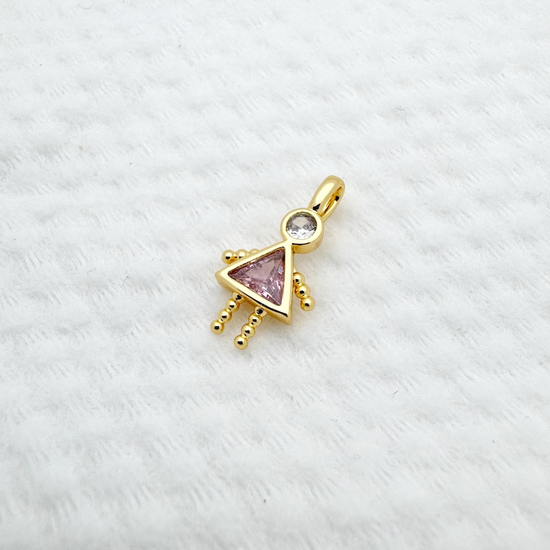 May include: A gold-colored pendant in the shape of a stylized person. The pendant features a pink triangle for the body, a clear round stone for the head, and small gold beads for the arms and legs. The pendant has a loop at the top for a chain.