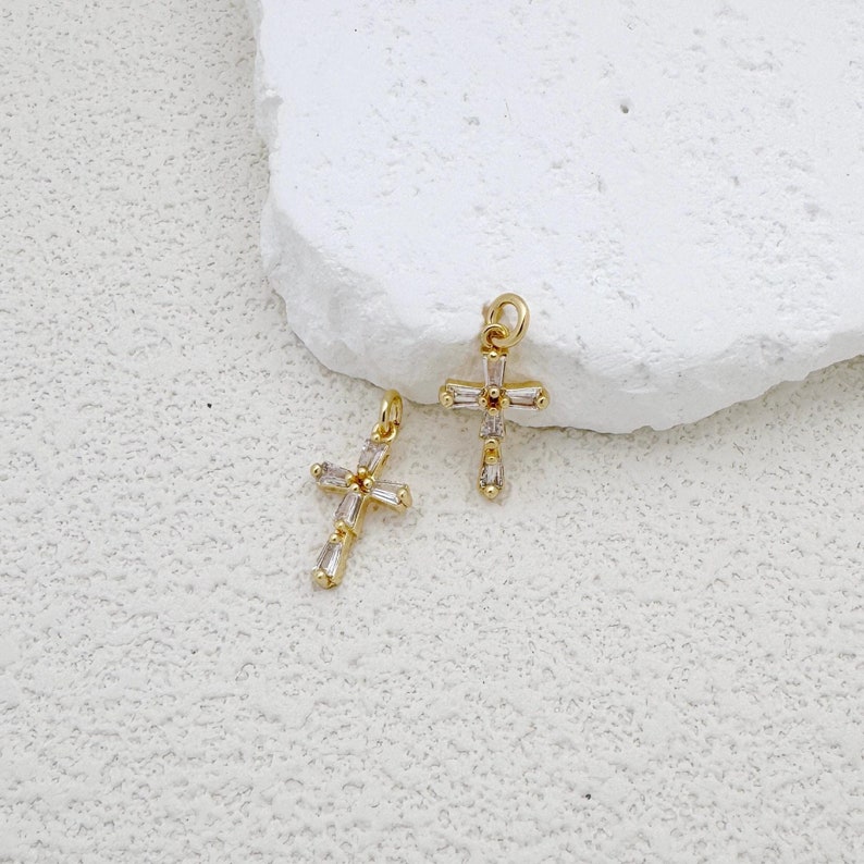 May include: Two gold cross charms with clear crystal accents. The charms are on a white background.