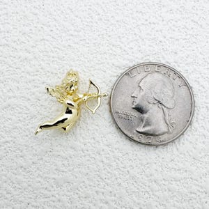 May include: A gold-colored cherub charm with a bow and arrow, next to a quarter coin. The cherub is in a flying pose, with detailed wings and curly hair. The coin shows a profile of a person and the words "IN GOD WE TRUST".