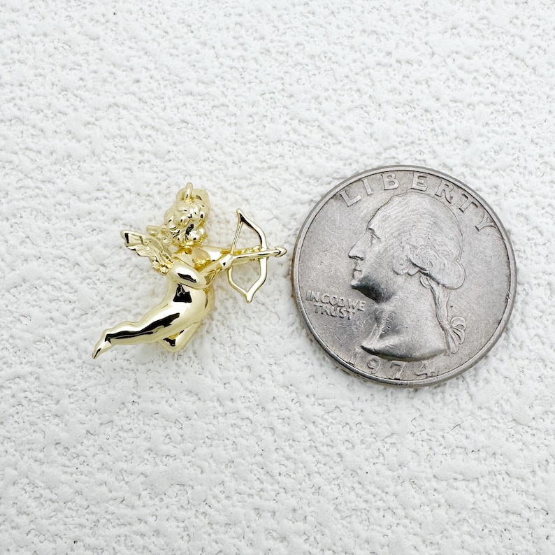 May include: A gold-colored cherub charm with a bow and arrow, next to a quarter coin. The cherub is in a flying pose, with detailed wings and curly hair. The coin shows a profile of a person and the words "IN GOD WE TRUST".