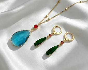 May include: A gold-tone necklace featuring a large, faceted, teardrop-shaped blue pendant and a small red circular accent. Two matching gold-tone hoop earrings each feature a slender, teardrop-shaped green pendant and a small red circular accent.