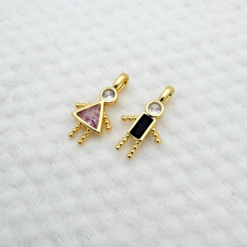 May include: Two gold-colored pendant charms in the shape of stick figures. One has a pink triangle body and a clear gem head, the other has a black rectangle body and a clear gem head. Both have small gold bead arms and legs.