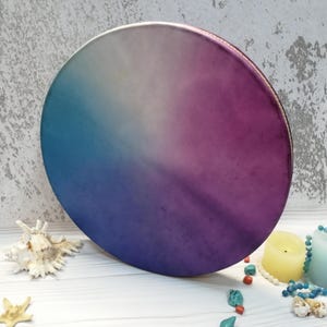 Puede incluir: Un tambor redondo pintado a mano con un diseño degradado azul, morado y blanco. El tambor está hecho de madera y tiene una cabeza de cuero.