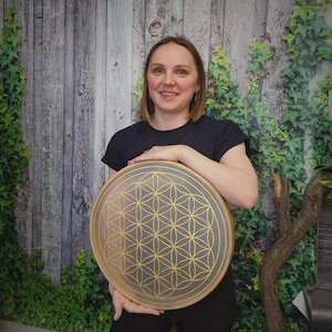 May include: A round, wooden drum with a gold-colored Flower of Life pattern. The drum is held by a person wearing a black shirt. The background features a wooden wall and green foliage.