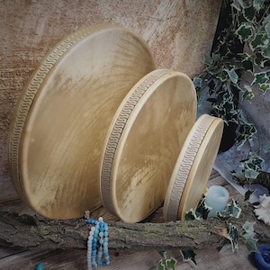 May include: Three wooden hand drums of different sizes with a decorative pattern around the edge. The drums are light brown and have a natural wood grain.