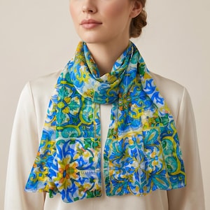 Può includere: Una sciarpa trasparente con un vivace motivo floreale nei toni del blu, giallo e verde. La sciarpa è drappeggiata intorno al collo di una persona, mostrando l'intricato design. La parola "MALIA" è stampata sulla sciarpa.