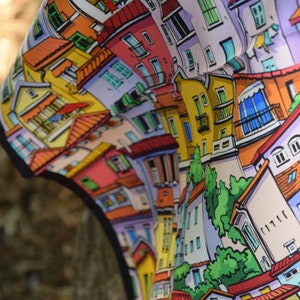 May include: A colorful scarf featuring a whimsical illustration of a cityscape with buildings in shades of yellow, pink, and blue. The scarf has a black border and a detailed design, perfect for adding a pop of color to any outfit.