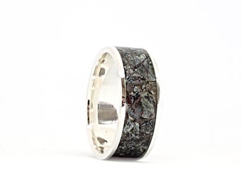 Puede incluir: Un anillo de plata con una banda ancha con una incrustación texturizada de color verde oscuro y gris. El anillo tiene una superficie pulida y reflectante y un diseño moderno. La incrustación parece estar hecha de piedra o material compuesto.