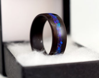 May include: A dark wooden ring with a vibrant blue and purple inlay. The ring is set in a black box with white padding, highlighting the contrast between the dark wood and the colorful inlay. The ring's design is simple yet elegant.