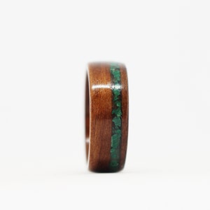 May include: A wooden ring with a polished, dark brown finish. A band of small, irregular green stones is inlaid around the center of the ring. The ring is set against a white background.