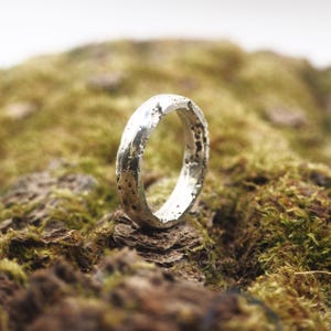 May include: A silver ring with a textured surface, set against a backdrop of green moss and brown bark. The ring's design features an uneven, organic texture, giving it a rustic appearance. The ring is a piece of jewelry.