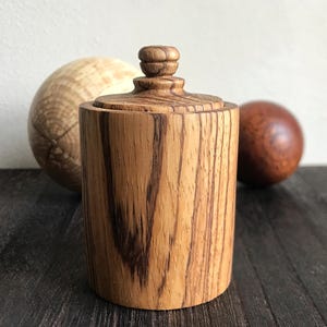 Puede incluir: Un recipiente de madera con tapa, hecho de madera con veta marrón oscura. El recipiente es cilíndrico y tiene un acabado liso y pulido.