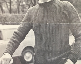 Può includere: Immagine in bianco e nero di un uomo che indossa un maglione a coste grigio scuro con una camicia bianca con colletto sotto. Il maglione ha uno scollo alto e rotondo. È visibile anche il testo "PASSAT DUOMATIC P.12. 9p 1081".