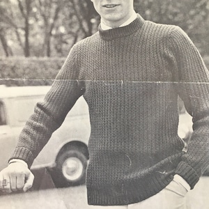 May include: A black and white image of a man wearing a dark gray ribbed knit sweater with a white collared shirt underneath. The sweater has a high, round neckline. The image also includes text that reads "PASSAT DUOMATIC P.12. 9p 1081".