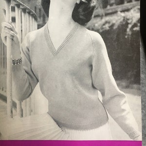 May include: A black and white image of a woman wearing a light-colored V-neck sweater and a headband. The number "253" is in a circle in the upper right corner. The woman is looking up and smiling.