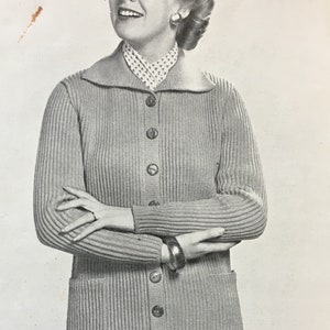 May include: A close-up of a woman wearing a grey knitted cardigan with a collared neckline and button fastening. The cardigan has two pockets on the front and is ribbed throughout. The woman is wearing a white blouse with a small pattern underneath the cardigan.