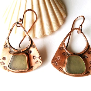 May include: A pair of copper earrings featuring a fan-shaped metal base with stamped floral patterns and a central piece of frosted sea glass. The earrings are suspended from curved metal ear wires, showcasing a warm metallic finish.