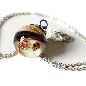 May include: A clear resin pendant necklace with gold leaf flakes and dark brown hair inclusions. The pendant is spherical and hangs from a silver chain. The necklace is displayed on a white background.