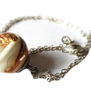 May include: A silver chain necklace with a clear, spherical pendant. The pendant contains gold leaf and a cream-colored swirl design. The necklace is displayed against a white background, highlighting the jewelry's details.