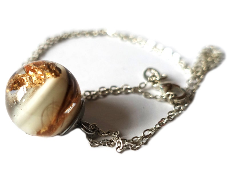 May include: A silver chain necklace with a clear, spherical pendant. The pendant contains gold leaf and a cream-colored swirl design. The necklace is displayed against a white background, highlighting the jewelry's details.