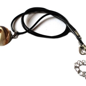 May include: A pendant necklace featuring a round, clear resin orb with gold flecks and brown and white swirls. The pendant hangs from a black cord with a silver-toned clasp and chain extender. The necklace is displayed against a white background.