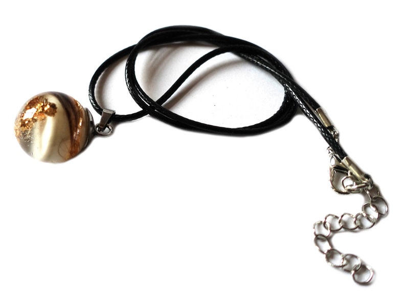 May include: A pendant necklace featuring a round, clear resin orb with gold flecks and brown and white swirls. The pendant hangs from a black cord with a silver-toned clasp and chain extender. The necklace is displayed against a white background.