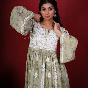May include: A green cotton dress with white embroidery. The dress has a V-neckline and long sleeves with bell cuffs. The dress is gathered at the waist and has a flowing skirt. The dress is photographed against a red background. &copy; The Chikankari Label.