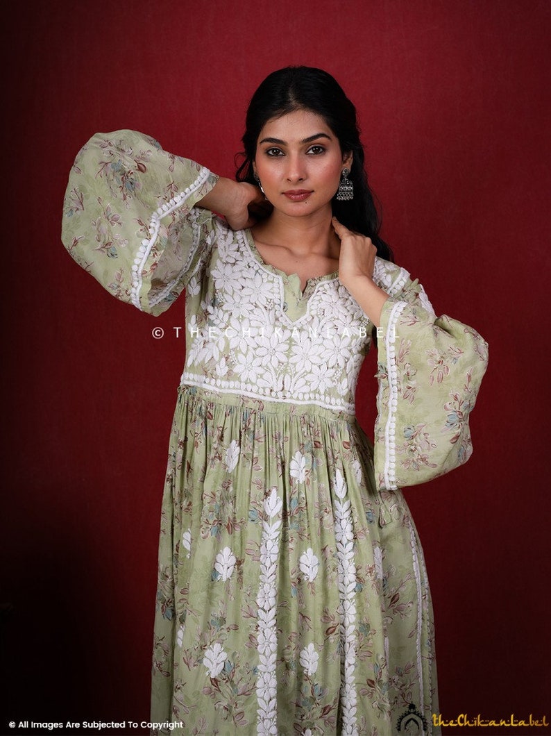 May include: A green cotton dress with white embroidery. The dress has a V-neckline and long sleeves with bell cuffs. The dress is gathered at the waist and has a flowing skirt. The dress is photographed against a red background. &copy; The Chikankari Label.
