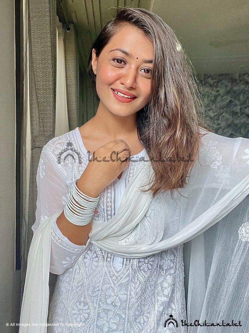 May include: A woman wearing a white embroidered kurta with a white dupatta draped over her shoulders. She is wearing silver bangles on her wrist. The text "thechikanlabel" is visible in the image.