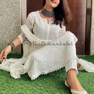 May include: A white chikankari kurta set featuring intricate embroidery and a delicate lace trim. The set includes a long-sleeved kurta, matching trousers, and a sheer dupatta. The model is wearing a silver necklace and bangles.